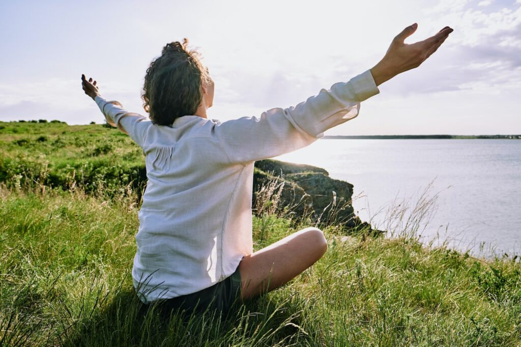 Happy young relaxed female with outstretched arms sitting on the ground by lake